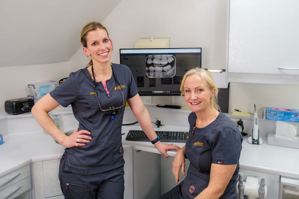 A portrait of the practice owners, Julia and Beverley, inside a dental surgery. Julia stands on the left wearing dark grey scrubs and dental loupes around her neck, smiling with her hand on her hip. Beverley sits on the right, also smiling and wearing matching dark grey scrubs. A computer screen displaying dental X-rays is visible behind them.