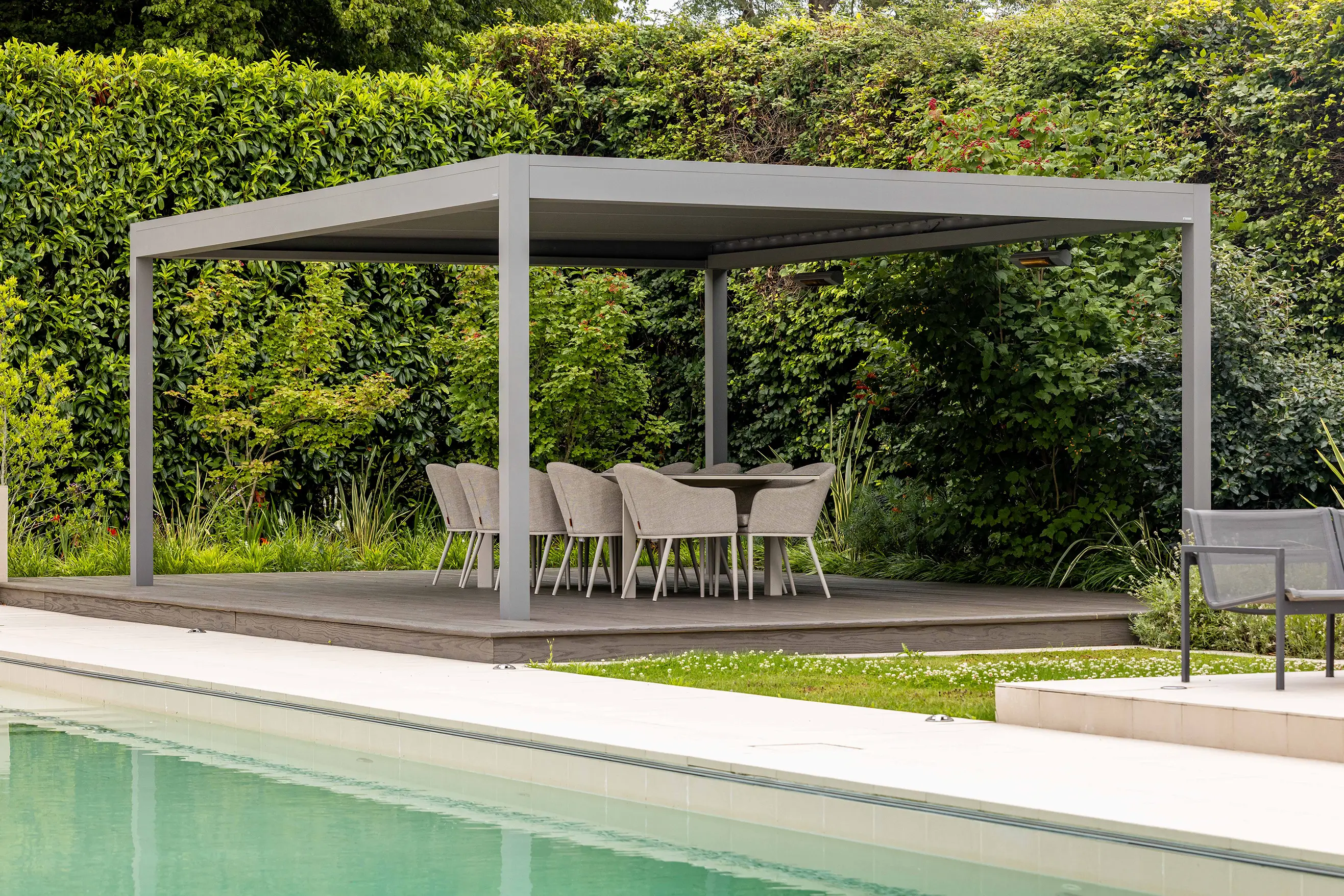 A modern pergola with seating beside a swimming pool, surrounded by lush green plants.
