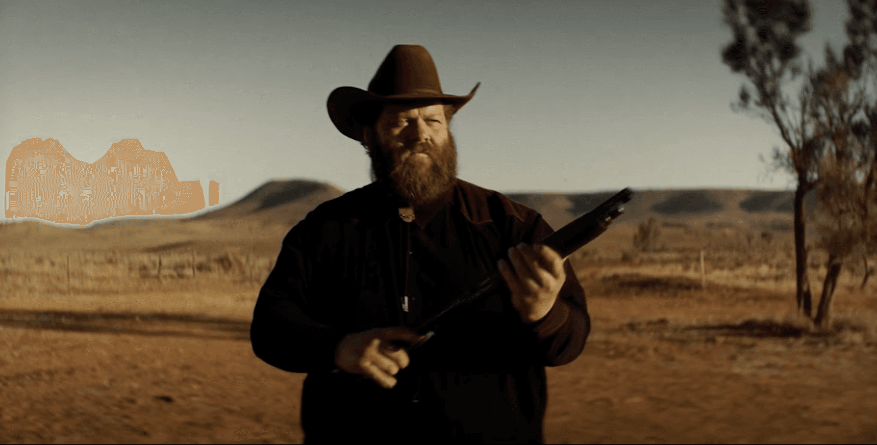 A man in a cowboy hat holds a weapon, standing in a desert landscape with mountains in the background.