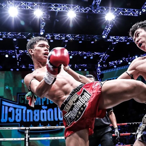 两名拳击手在拳台上，其中一名正踢向另一名。背景中可以看到明亮的灯光和显示屏。