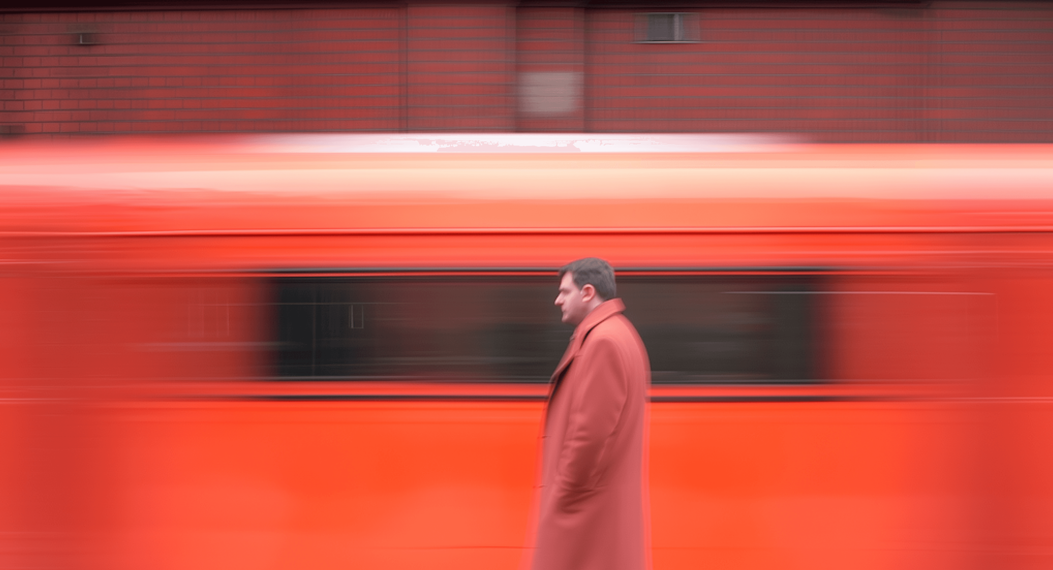 Man in red coat walks as a red vehicle speeds by, creating motion blur effect.