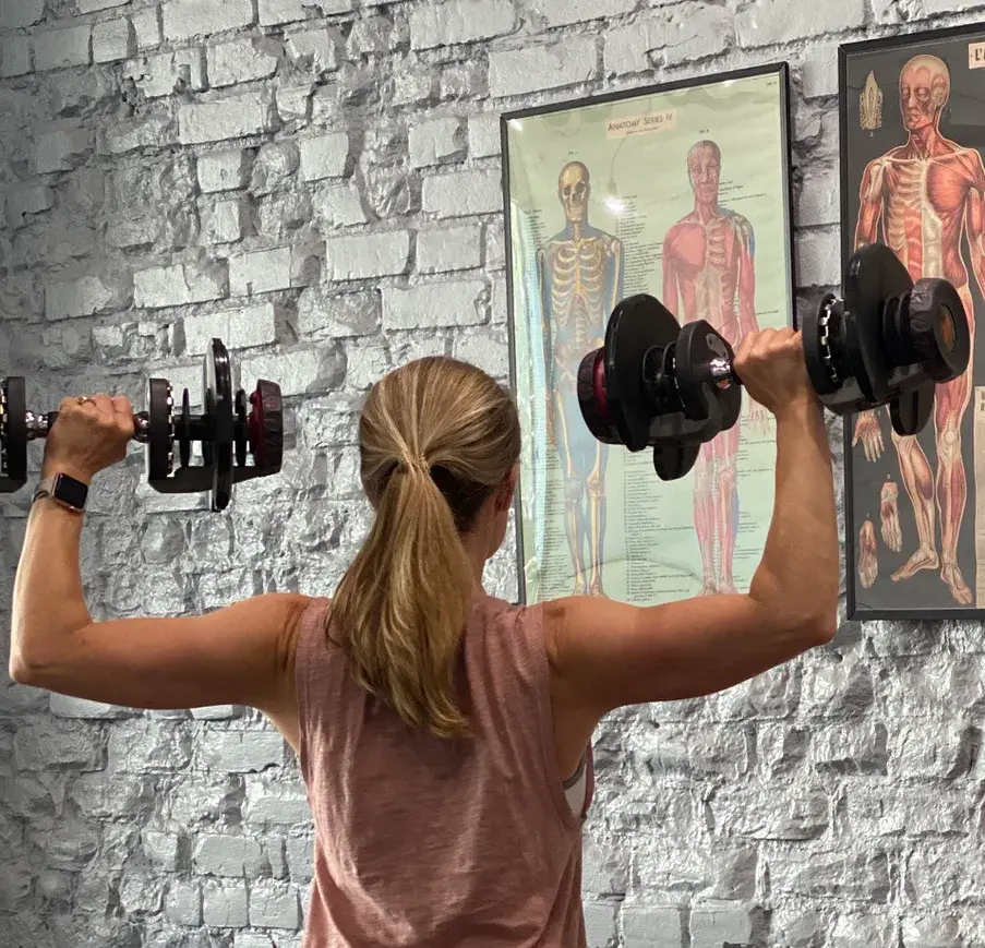 Heather Hausman in athletic clothing lifts adjustable dumbbells in front of a textured brick wall with anatomical posters, showcasing a fitness and anatomy theme.
