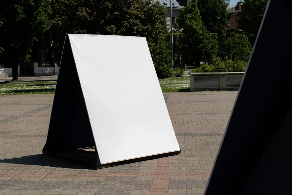 A-frame outdoor display with a blank white surface set in a sunny park environment. Surrounding elements include trees, pathways, and urban architectural details in the background.