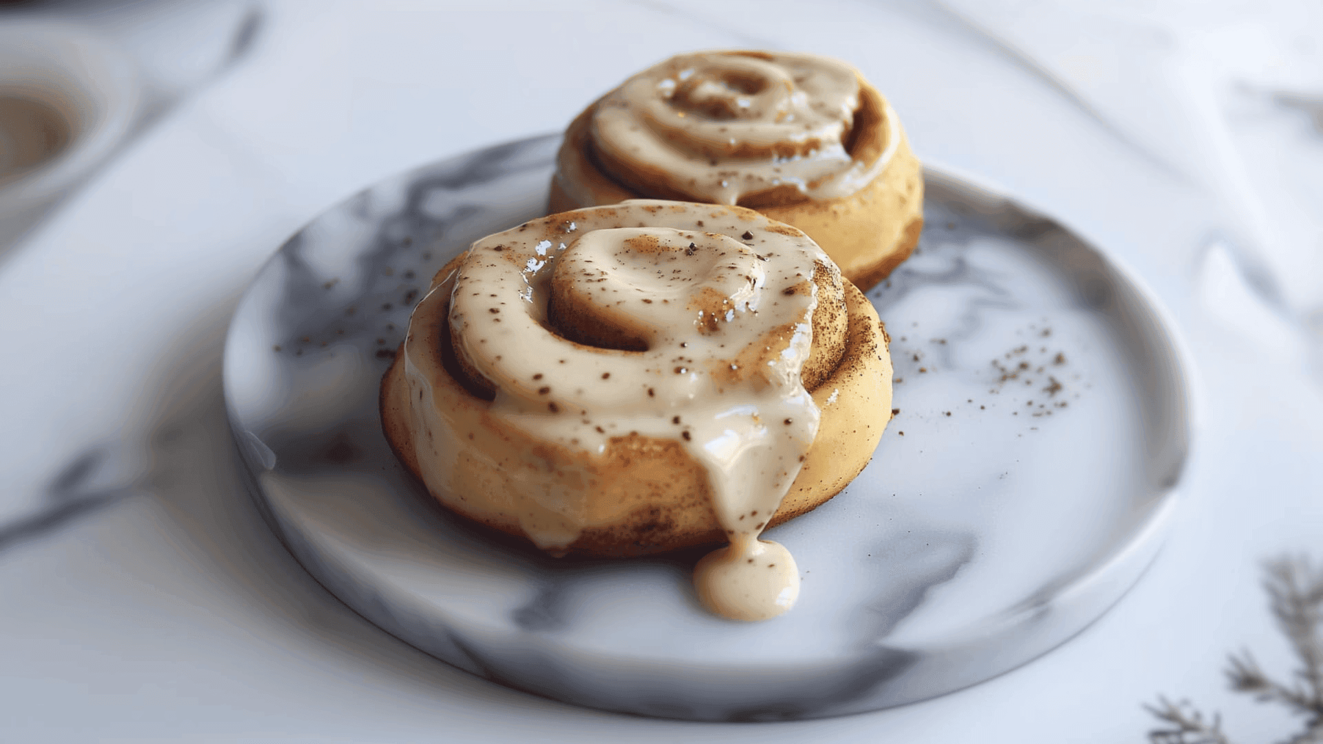 Zwei Zimtschnecken mit Protein-Frosting auf einem marmorierten Teller, weihnachtliche Deko im Hintergrund