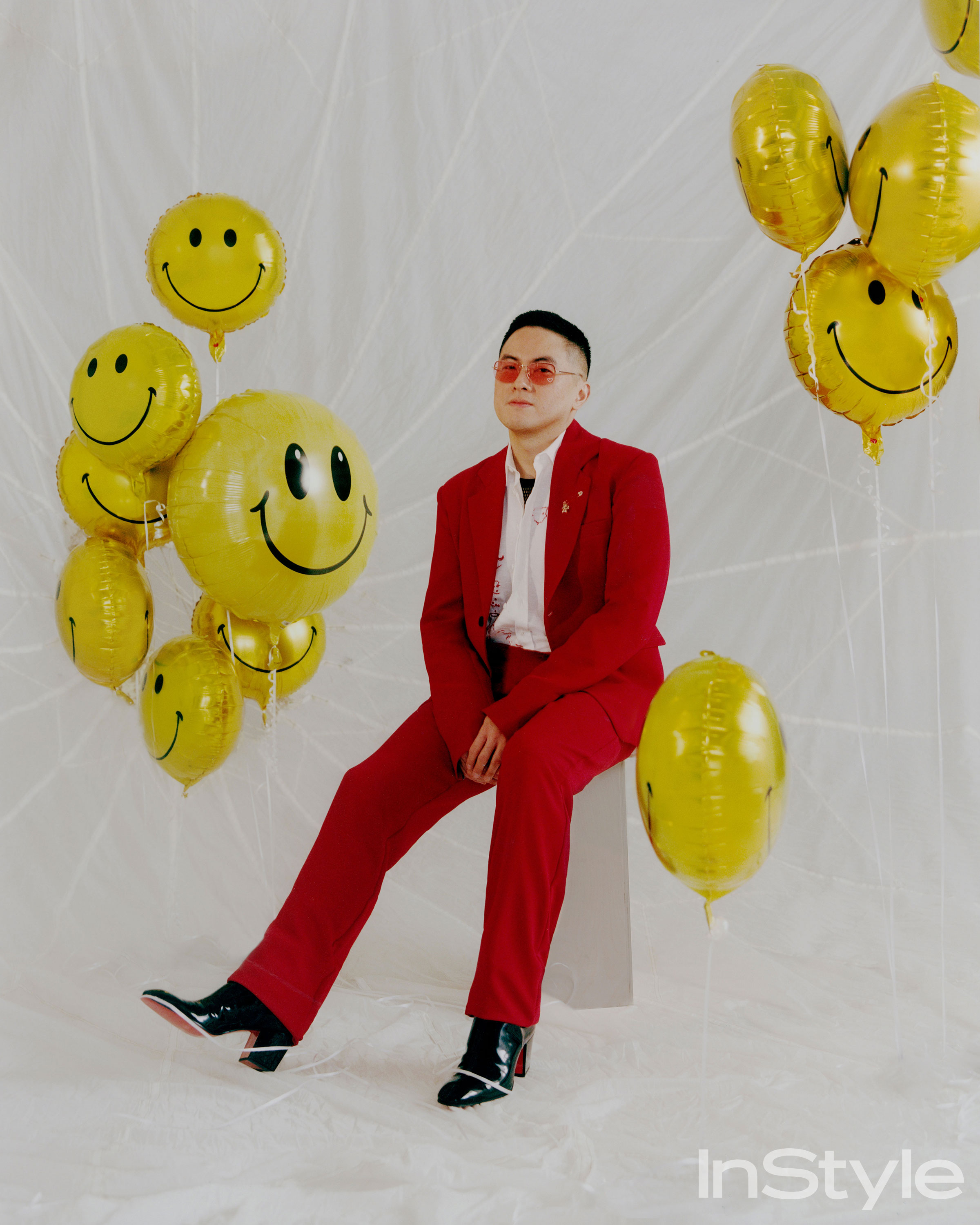 Bowen Yang in a red suit with gold smiling balloons for InStyle