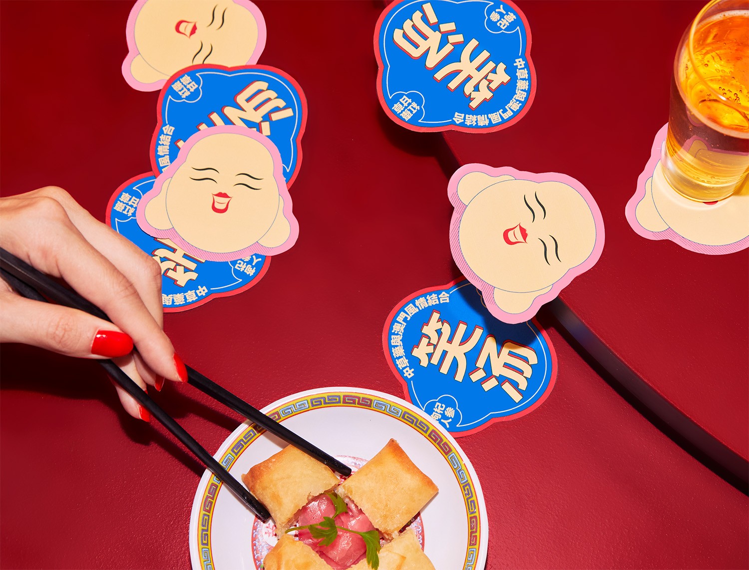 Macau Laughing Brew coasters with the smiling character illustration on a red table beside a plate and chopsticks.