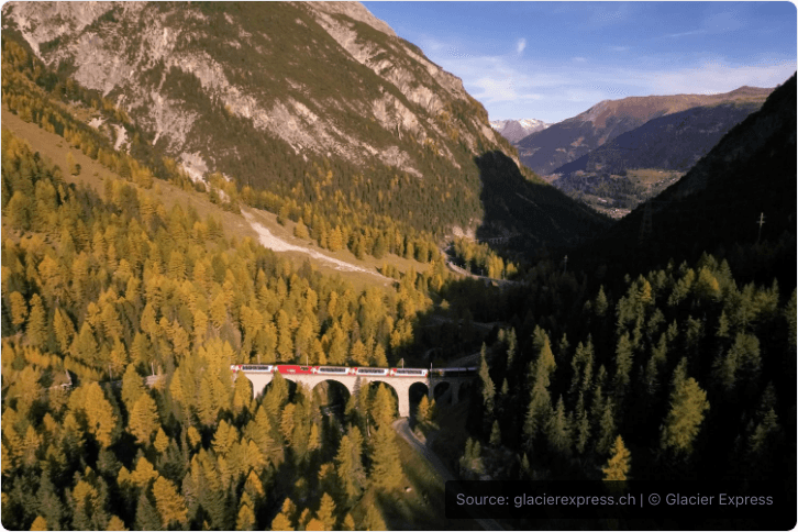 Glacier Express crossing a stone viaduct through autumn forest in the Swiss Alps.