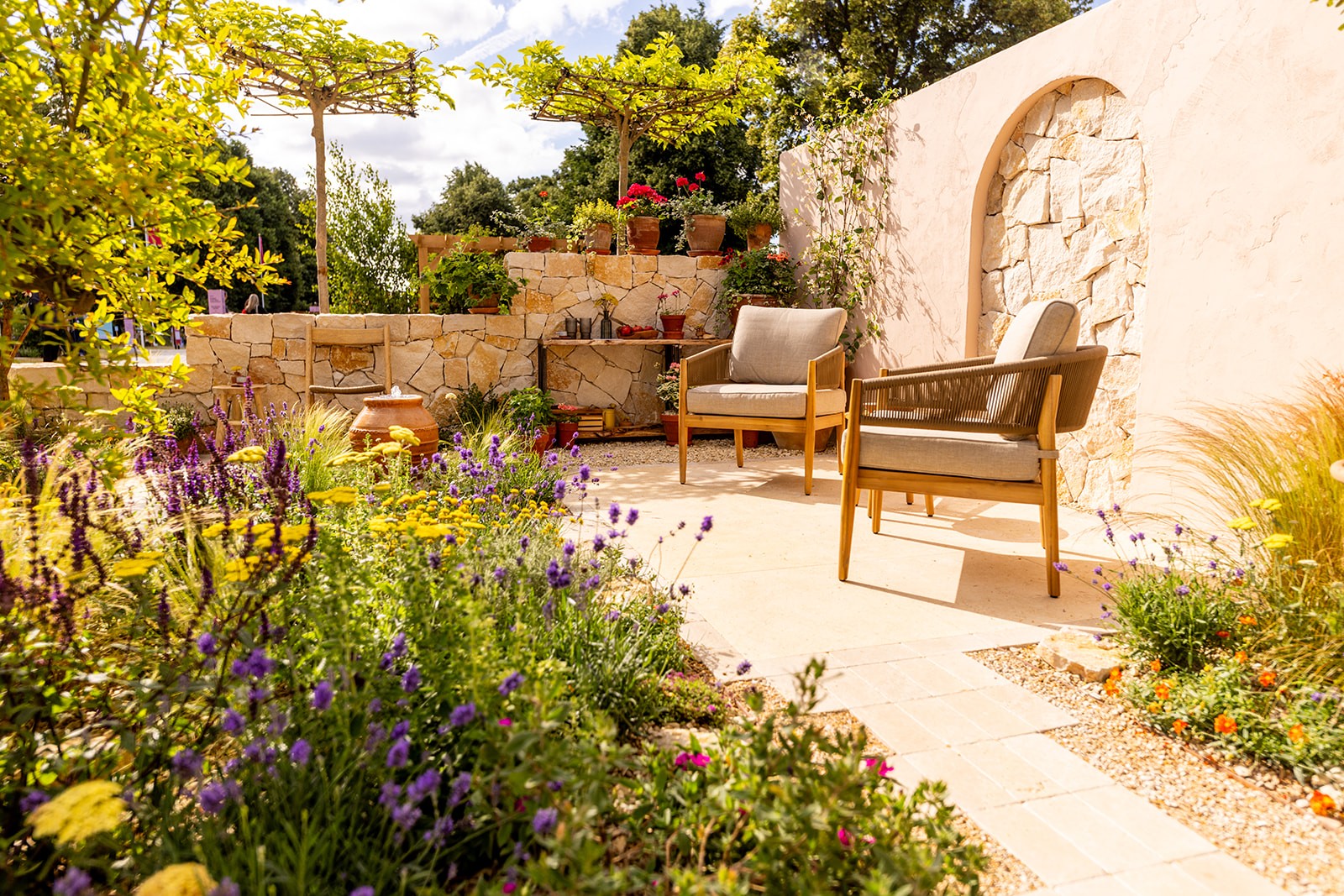 A serene garden patio with wooden furniture, surrounded by lush greenery and a stone wall. Bright and inviting atmosphere.