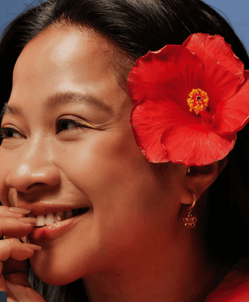A woman smiling with a large red hibiscus flower in her hair, wearing earrings decorated with floral designs.