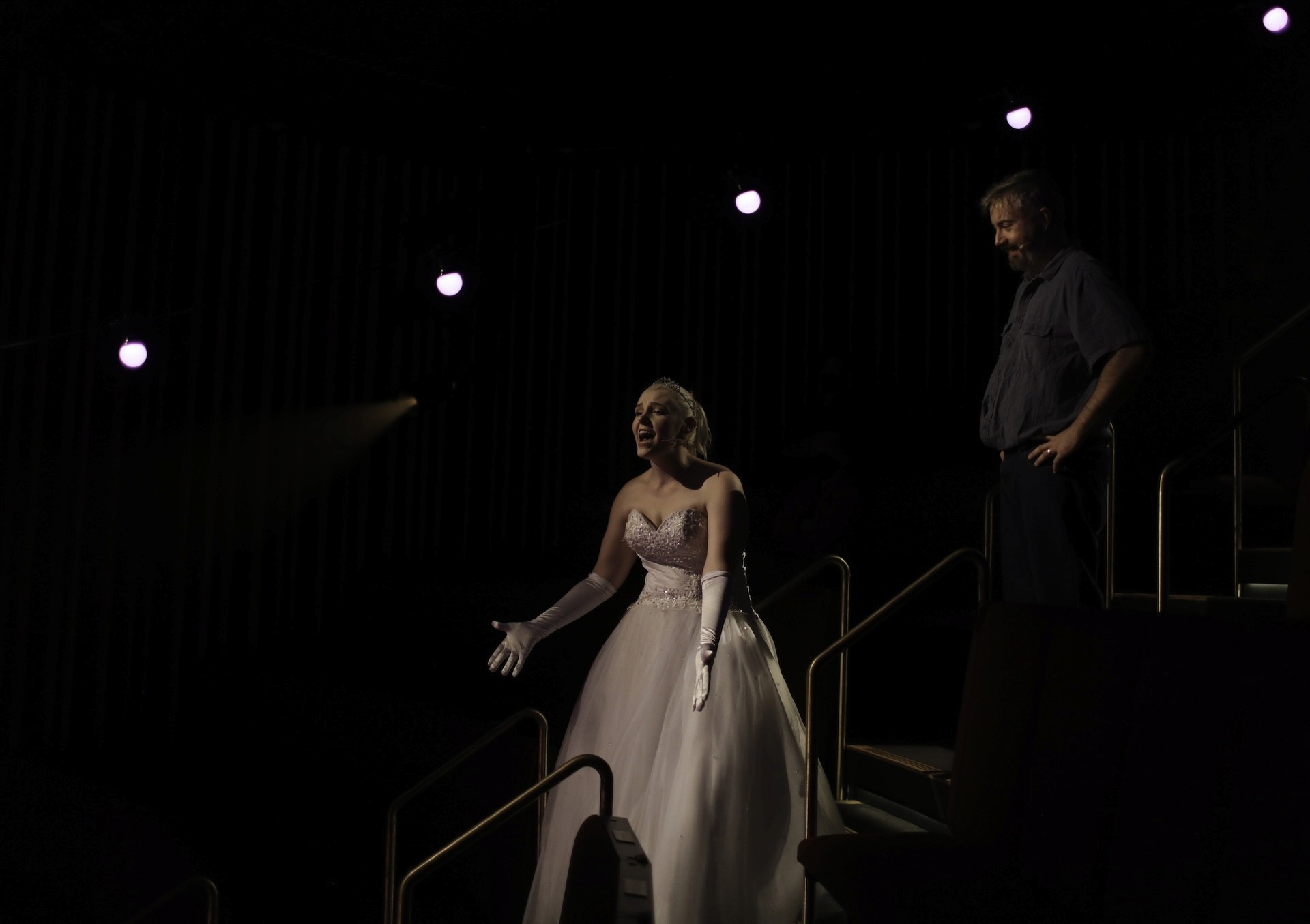 A performer in a white ballgown and long gloves stands in a spotlight on a theatre aisle, singing or speaking with outstretched hands. Another performer stands slightly behind her on the steps, watching quietly in the dimly lit auditorium.