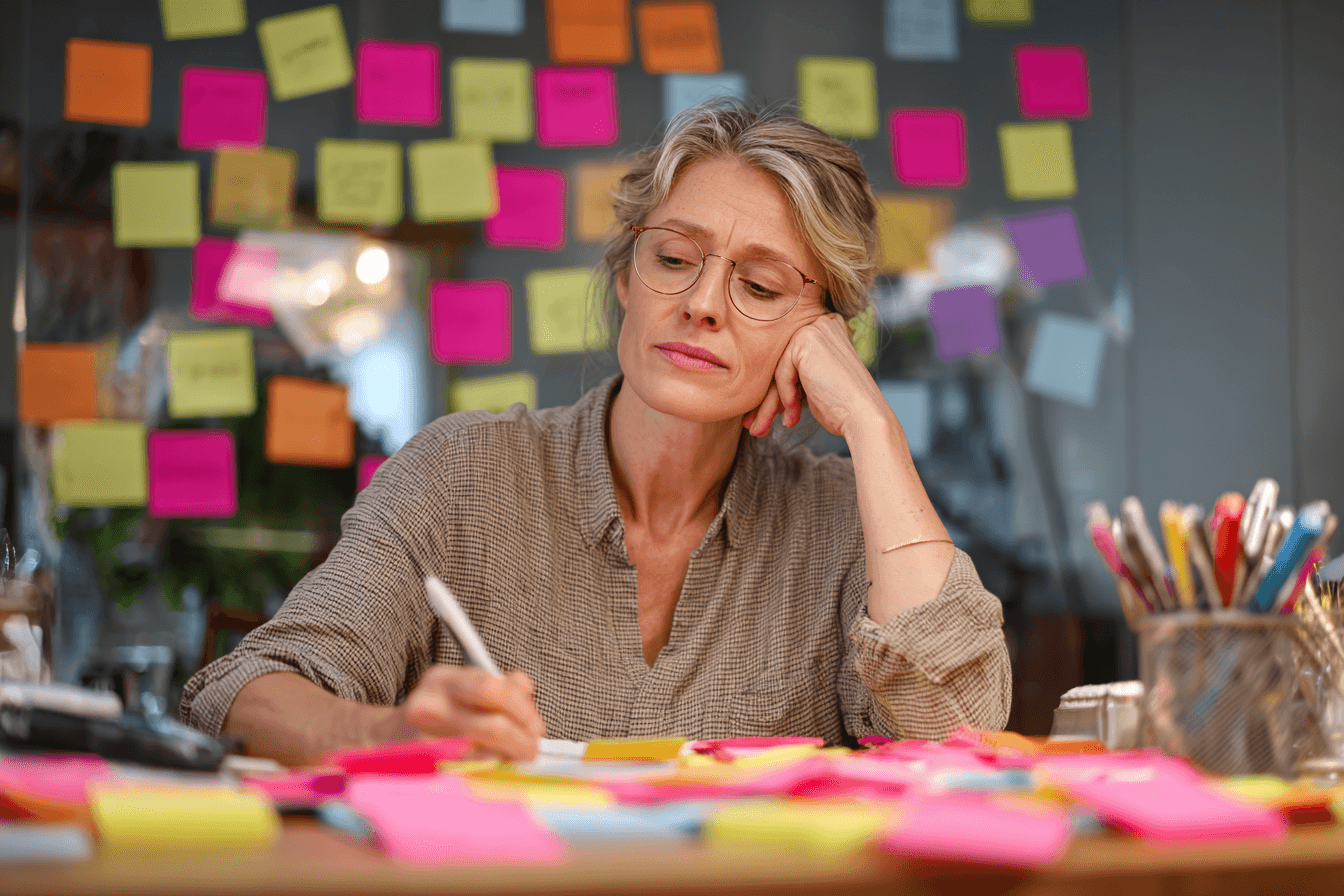 a home care agency marketer (female, 40s) in a home care agency office, sitting at her desk. She is writing on a sticky note. There are many sticky notes on her pinboard. She looks concerned.