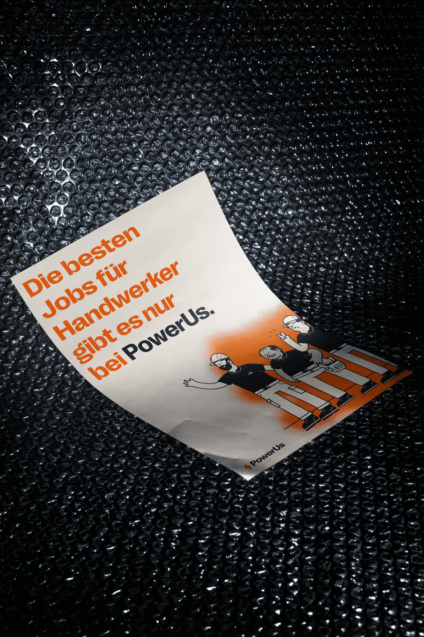 A textured surface holds a poster featuring cartoon construction workers and bold orange text promoting jobs for tradespeople with the PowerUs brand name.
