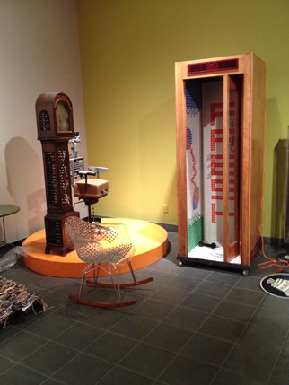 installation view of a sculpture made with a handbag and other metal pieces, and a tall wooden structure on the right side of the photo looks like a public phone cabin