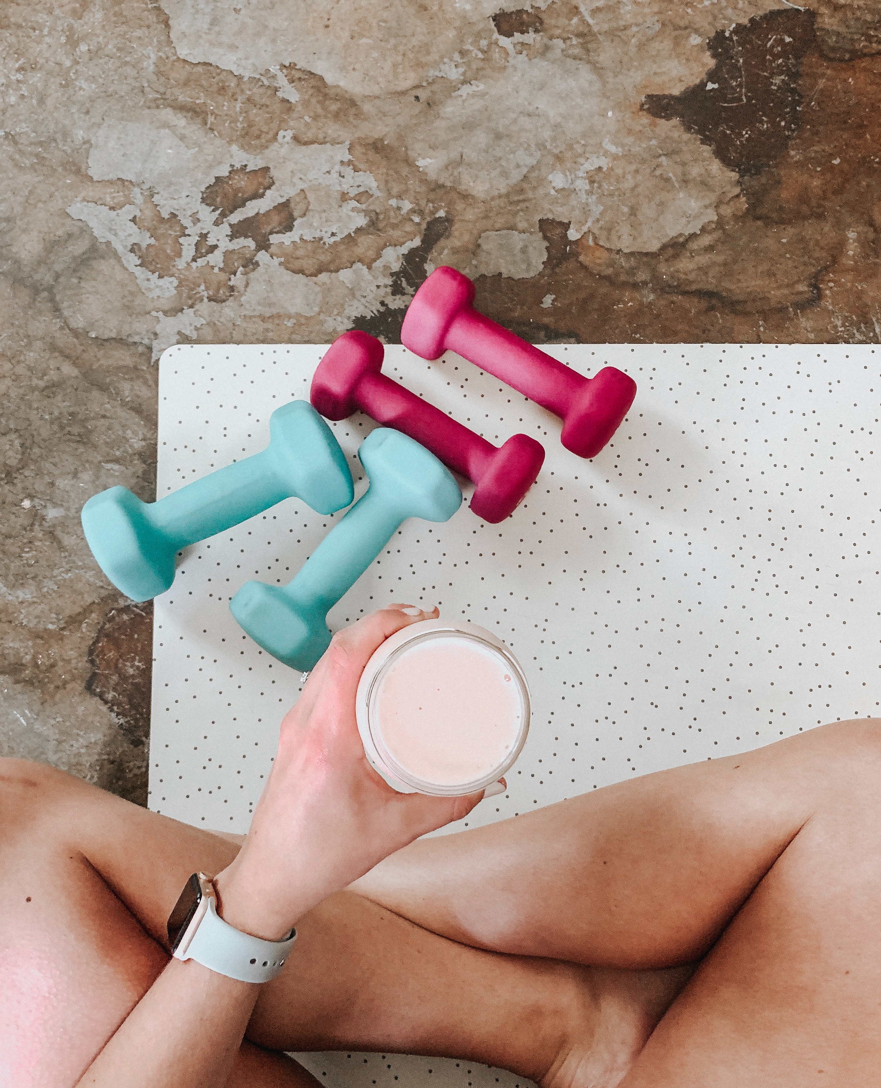 Woman sitting on a workout mat holding a protein shake, surrounded by light dumbbells—symbolizing recovery, home fitness, and strength training balance.