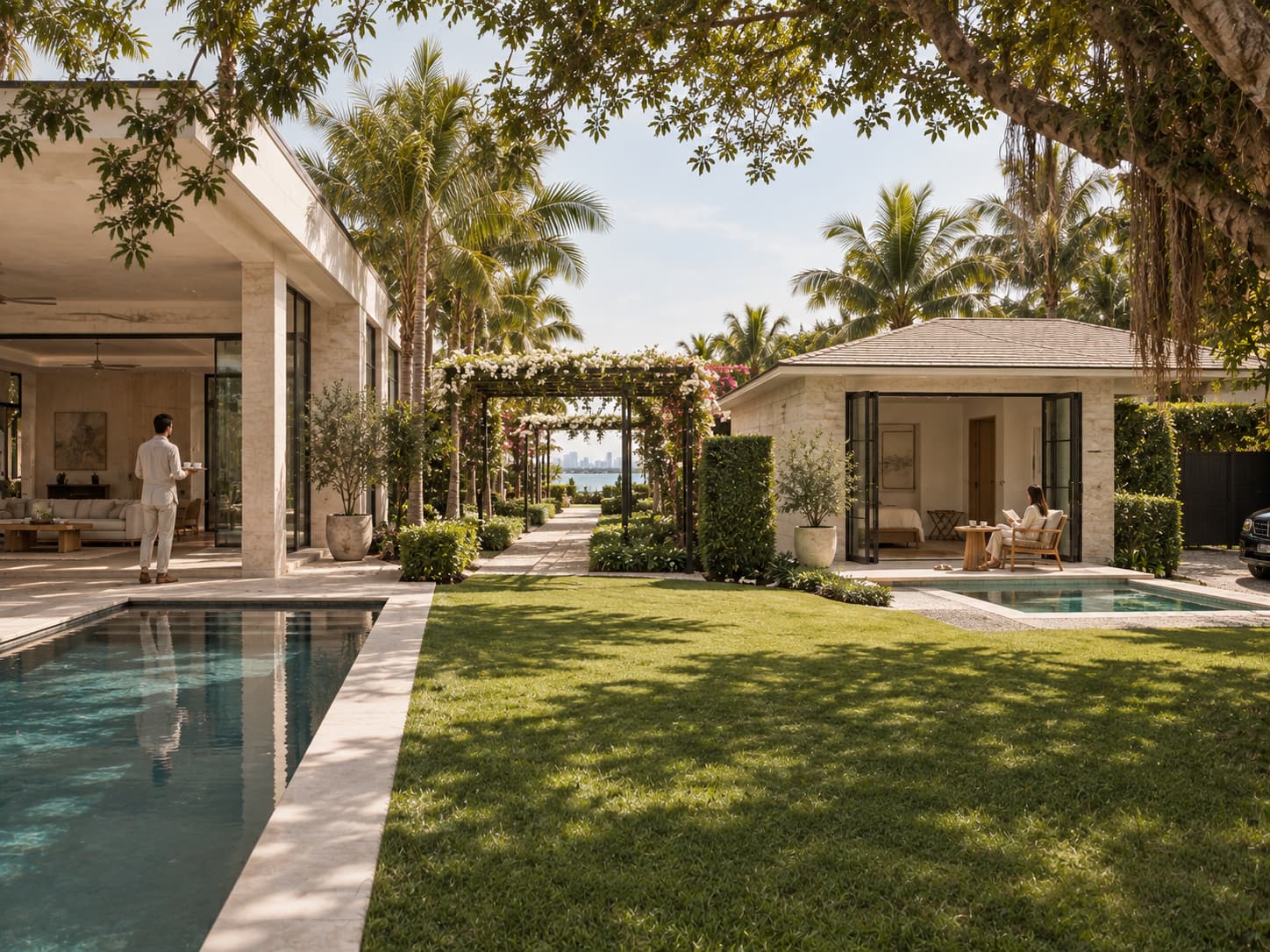 Sunlit estate garden with a lap pool, lawn and flowering pergola leading to the bay, where a man carries a tray on the main house terrace and a woman reads outside a guest cottage