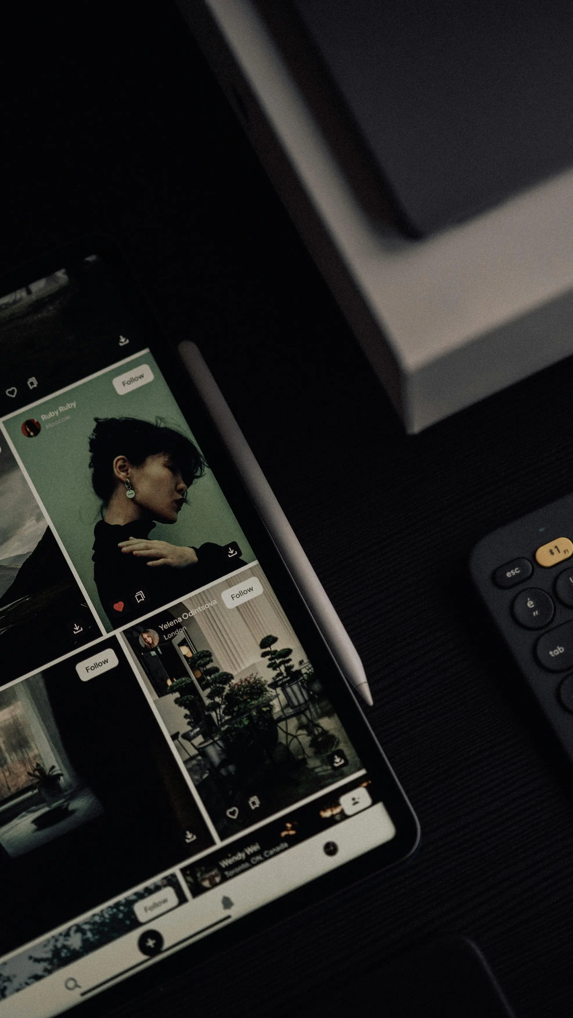 Tablet on a dark desk displaying a photo feed next to a stylus and keyboard. Moody, minimalist workspace.