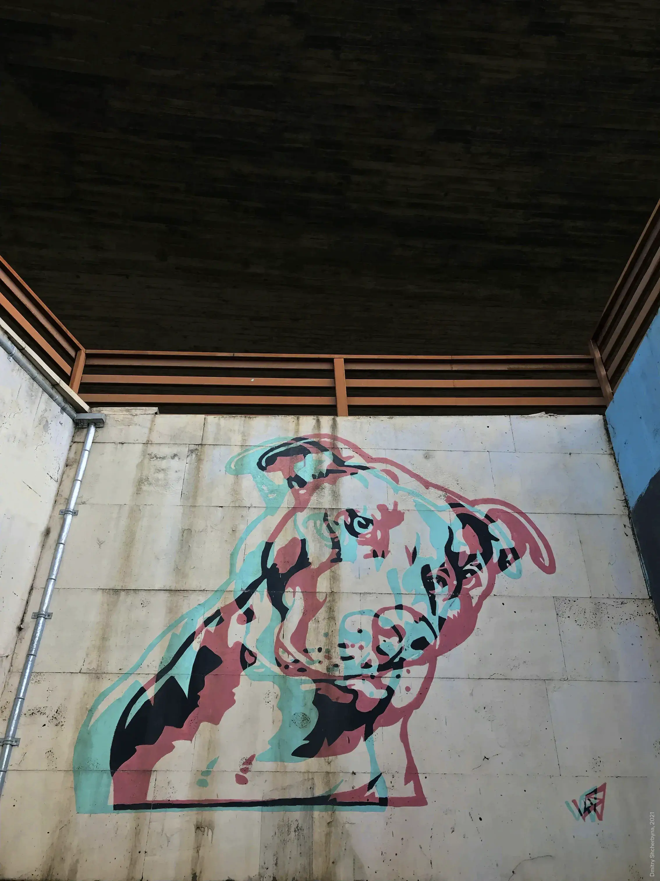 A large street mural of a pit bull, painted in pink, mint green and black, on a concrete wall beneath an overpass.