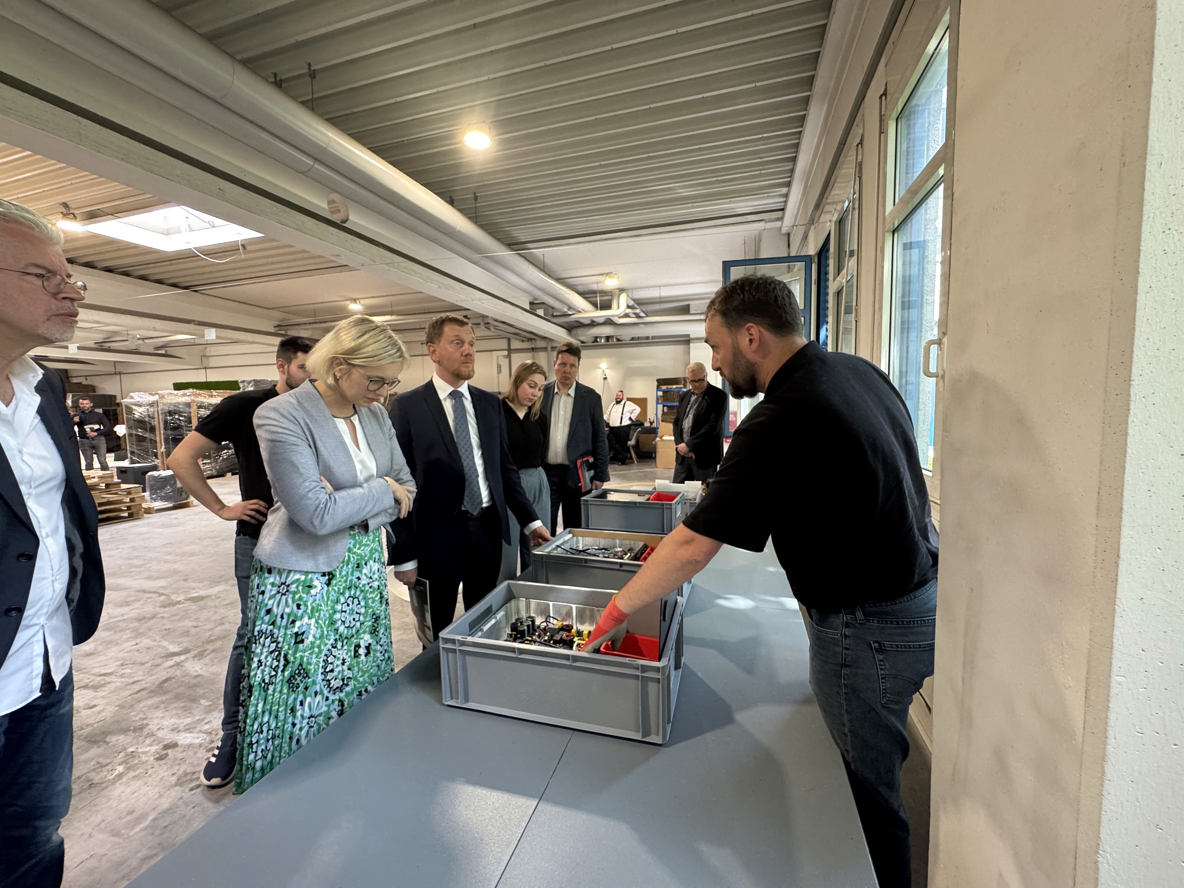 Besichtigung einer Fabrikhalle, Stefan Krüger, Ella Brimmers und Gruppe von Menschen beobachtet Arbeiter bei der Erklärung von Produktionsprozessen.