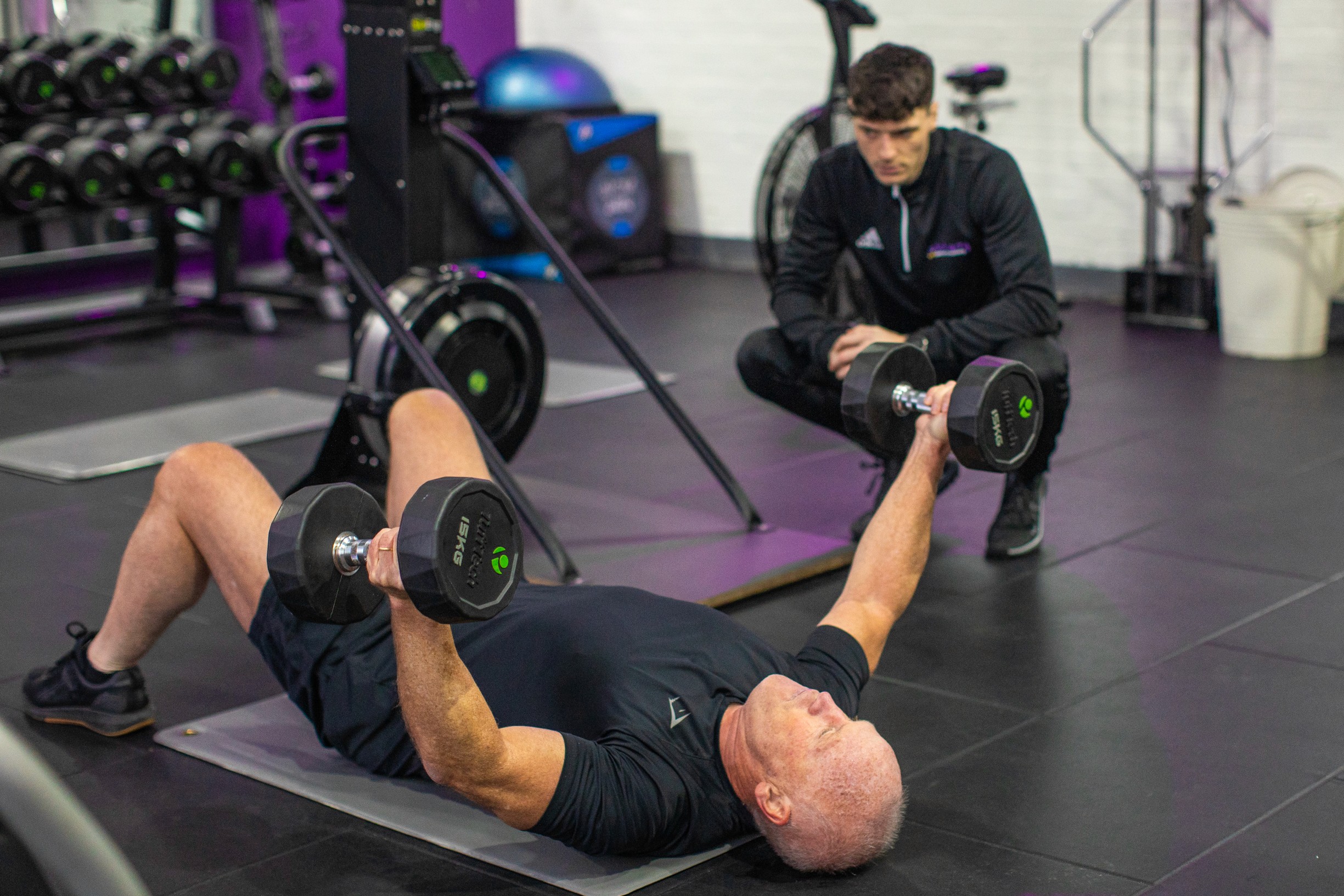 A man exercises on a mat with dumbbells in a gym. A trainer watches closely. The setting is modern, conveying focus and determination.
