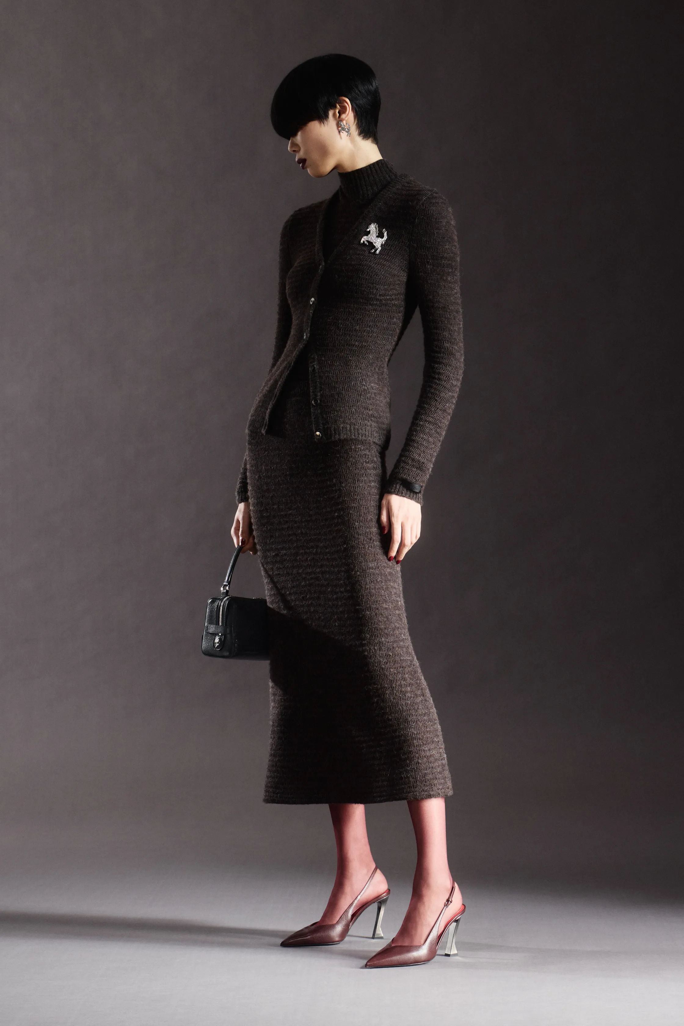 A model in a fitted dark knit top and matching midi skirt poses in a studio, holding a small handbag and wearing pointed heels for the Ferrari Pre-Fall 2026 collection.