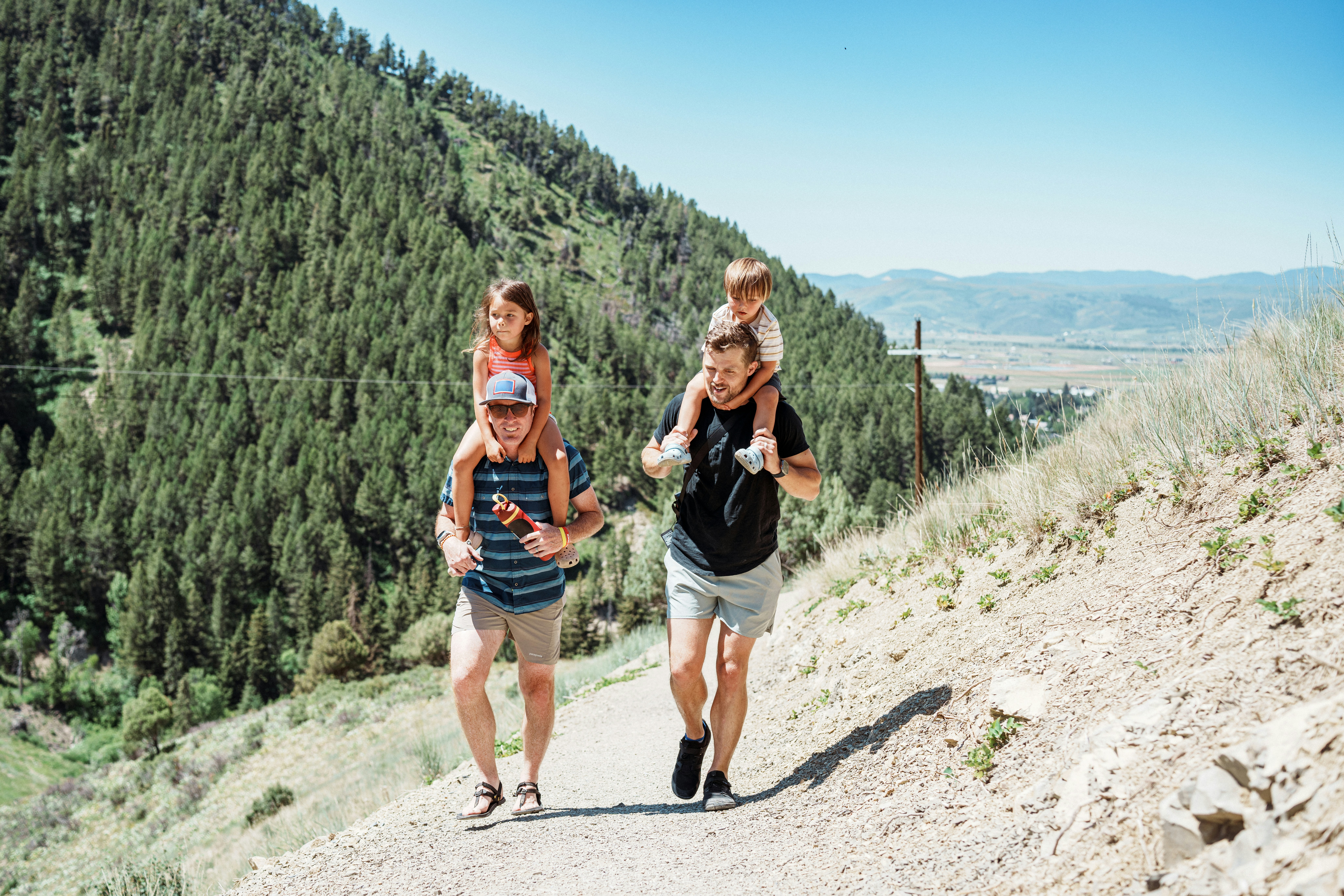 Two fathers carrying children on shoulders on a mountain path.