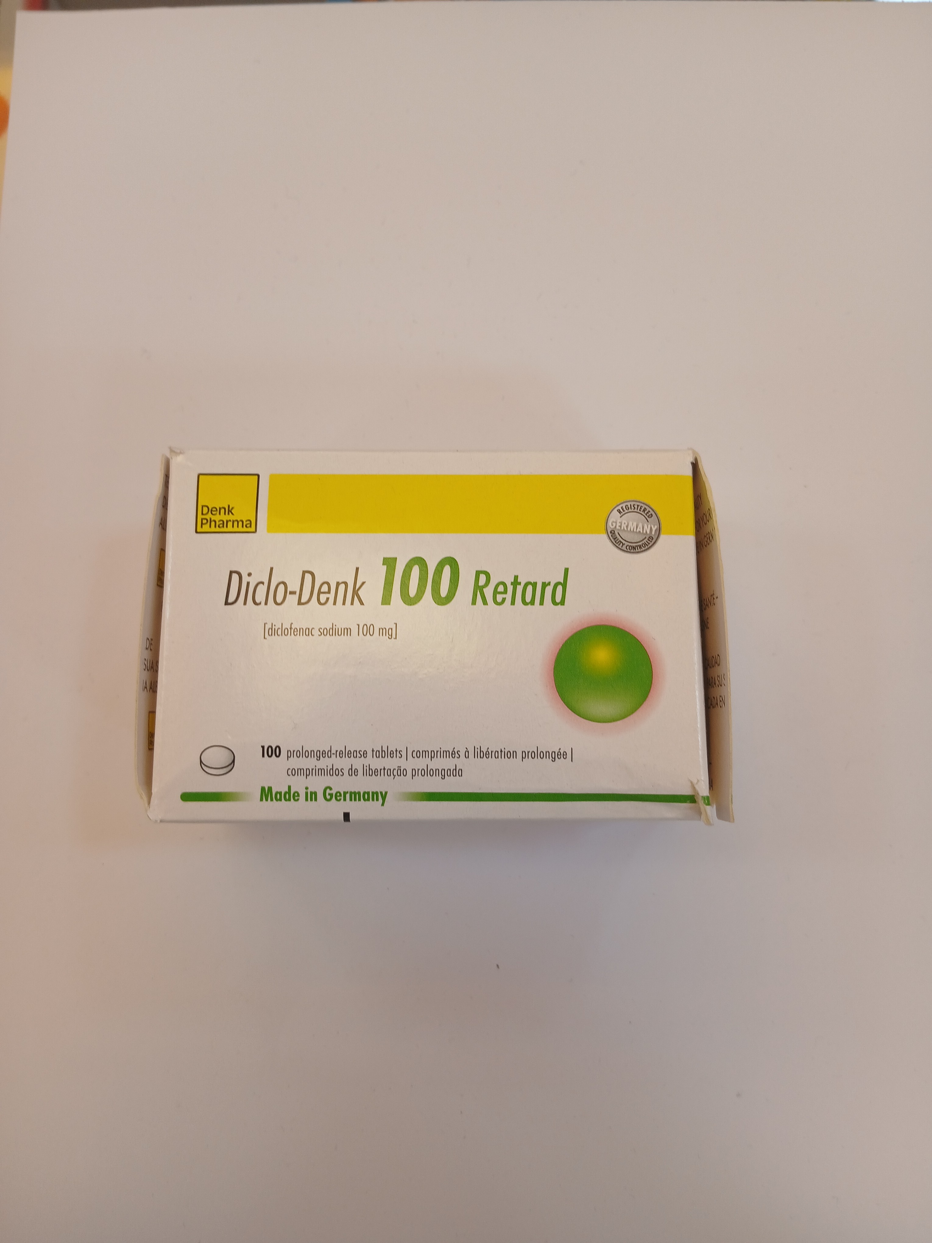 A small white cardboard box with a green and yellow label displays printed text in multiple languages and features a green circular pill inside a blister pack.