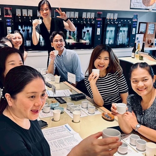 Un grupo de personas sonriendo y sosteniendo tazas, reunidas alrededor de una mesa con pequeñas tazas y papeles, en una habitación bien iluminada con un dispensador de bebidas.