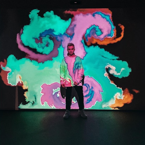 Person standing in front of a colorful swirling light projection, wearing casual clothing, in a dark room.