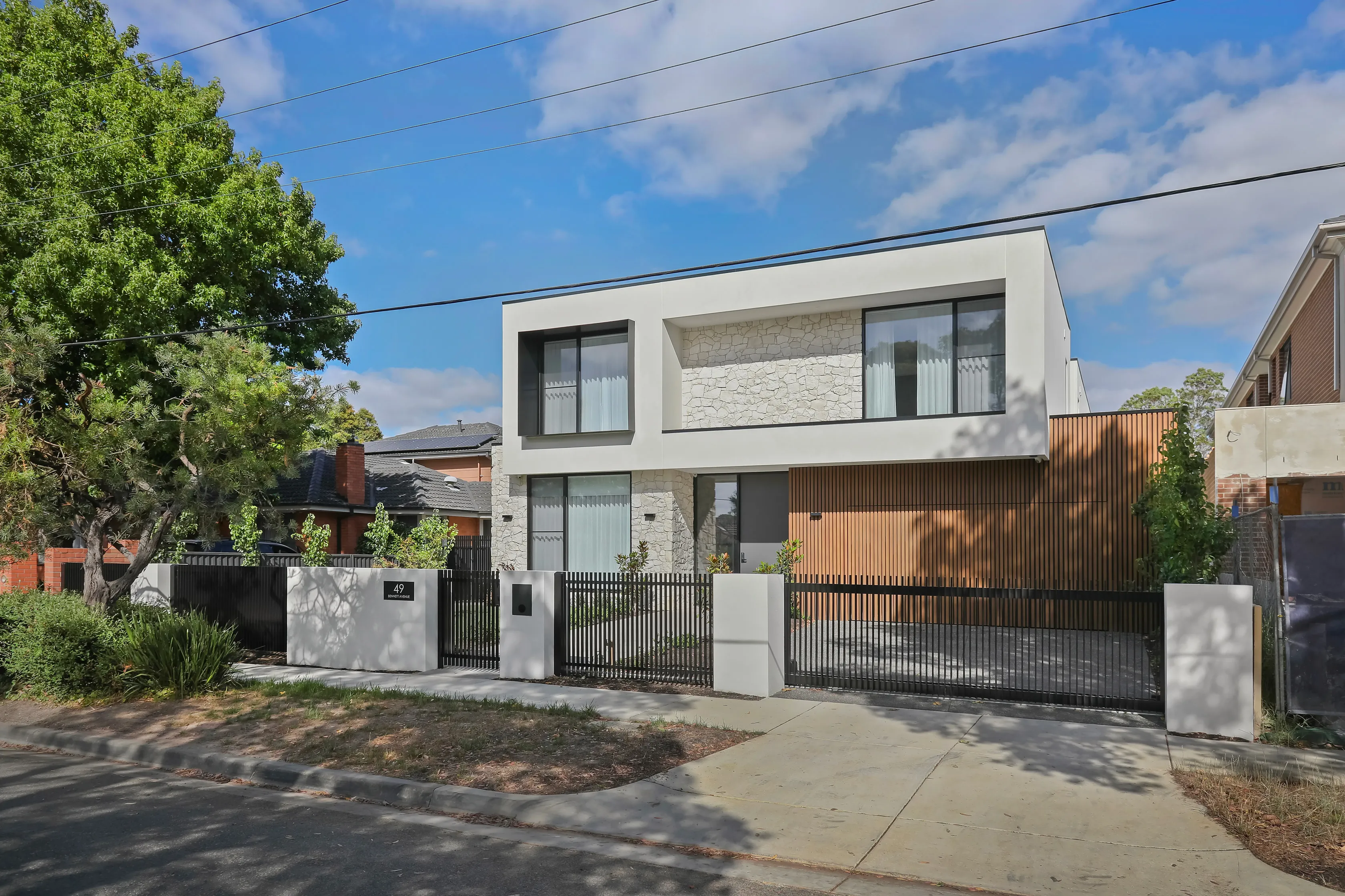 Street view of the Mount Waverley Residence, a modern two-storey home with white render, stone cladding and timber-batten facade behind a black picket fence