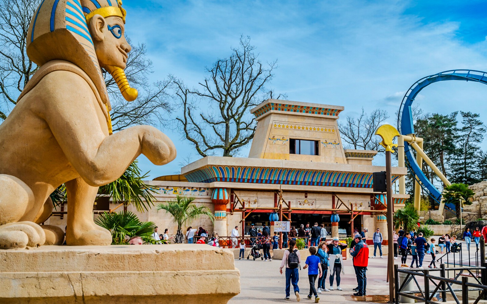 Roller coaster loop and Egyptian-themed entrance at Parc Asterix, France, with tourists exploring.