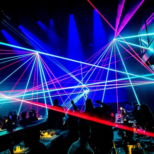 Orang-orang duduk di meja-meja dalam ruangan yang gelap, menyaksikan lampu laser warna-warni menciptakan pola di udara.