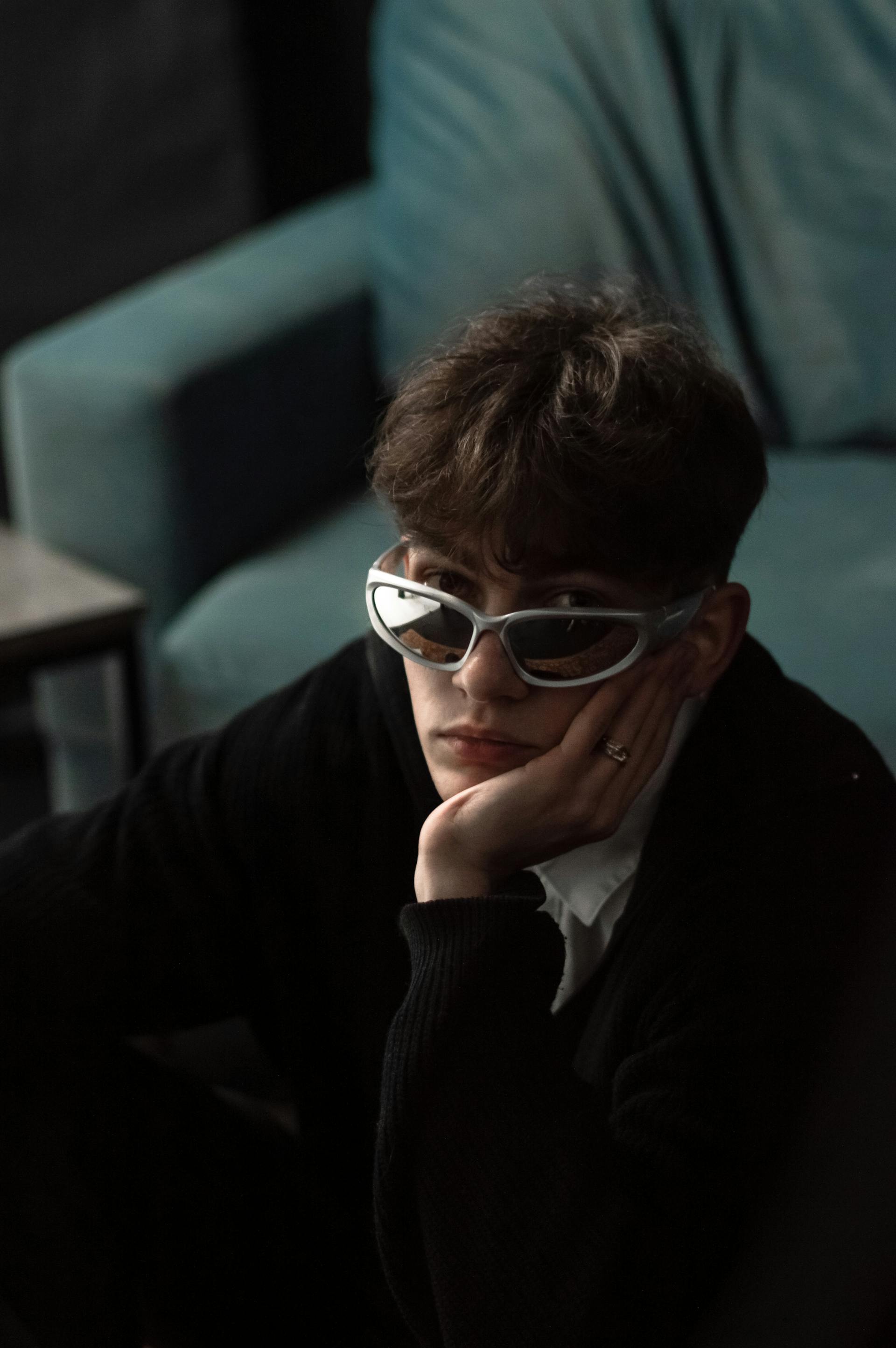 A young person with brown hair, wearing reflective silver sports sunglasses and a black sweater, looks at the camera while resting their chin on their hand.