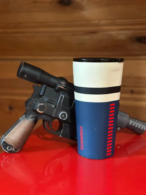 A Corkcicle tumbler with a minimalist Han Solo design in front of Han's trademark blaster. Both are sitting on a red surface with a wooden background