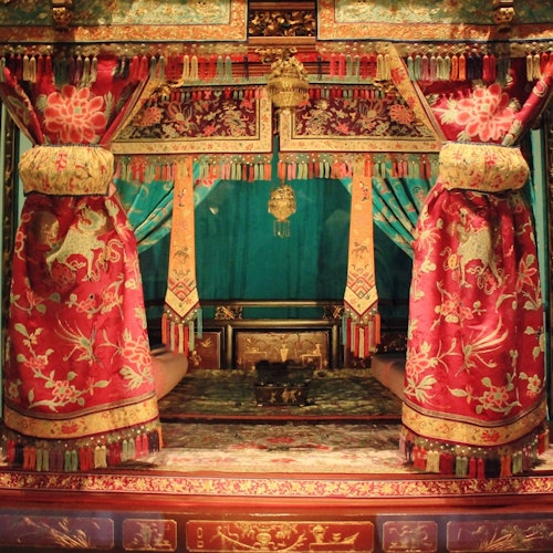 Ornate, colorful room with intricate red and gold drapes framing a cushioned seating area and decorative gold elements.