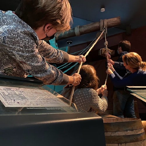 Four people pull ropes attached to a large wooden mechanism in an indoor museum exhibit.