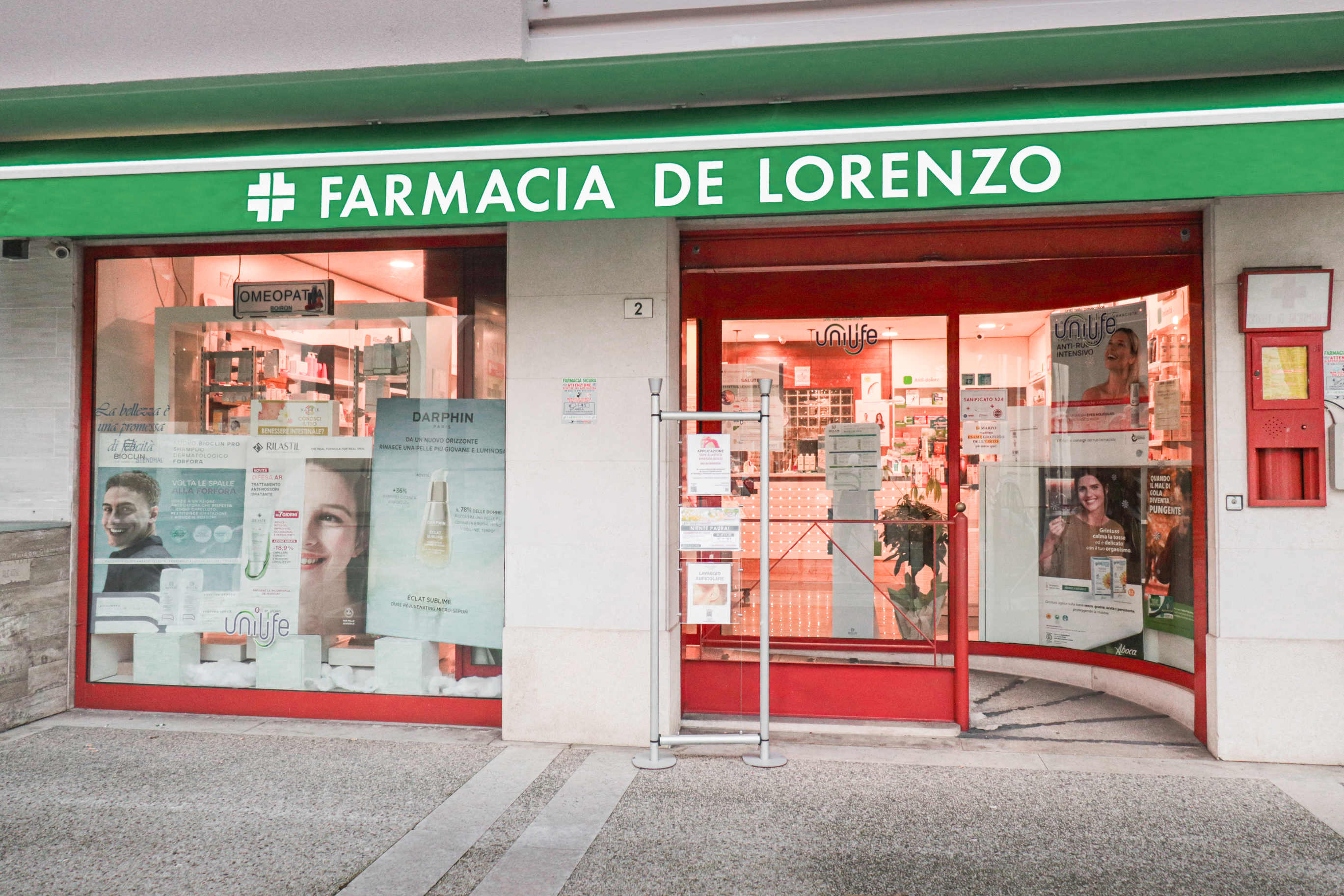 Vista dell'esterno della farmacia con le vetrate illuminate e il tendone marchiato.