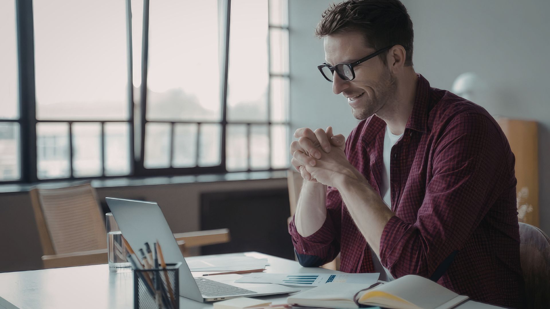 A man in glasses smiles at his laptop at a bright desk