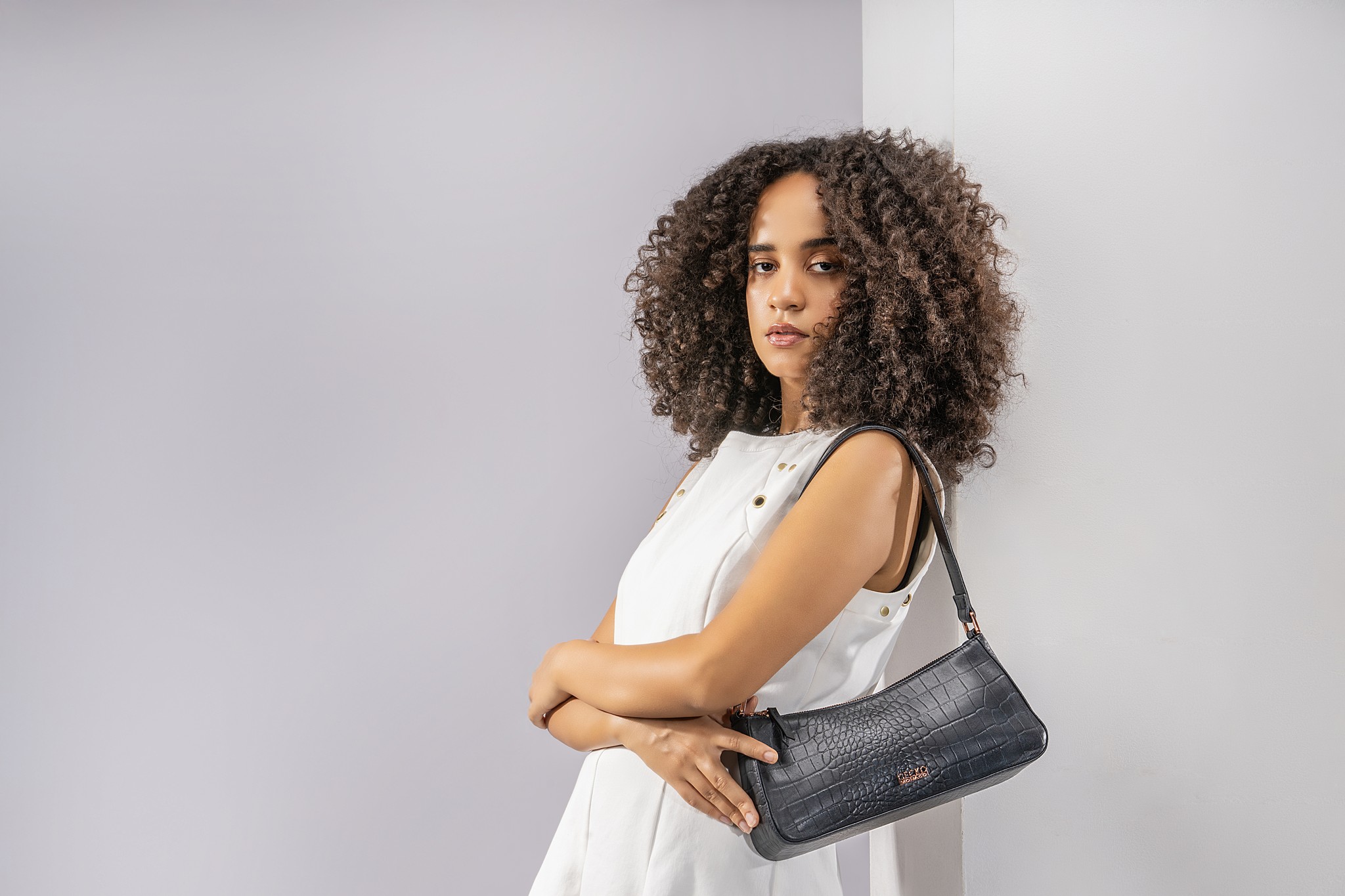 A woman with curly hair poses confidently against a light background, holding a stylish black handbag.