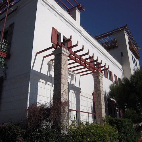 Large white building with stone columns and red wooden beams. Greenery and trees in the foreground against a clear blue sky.
