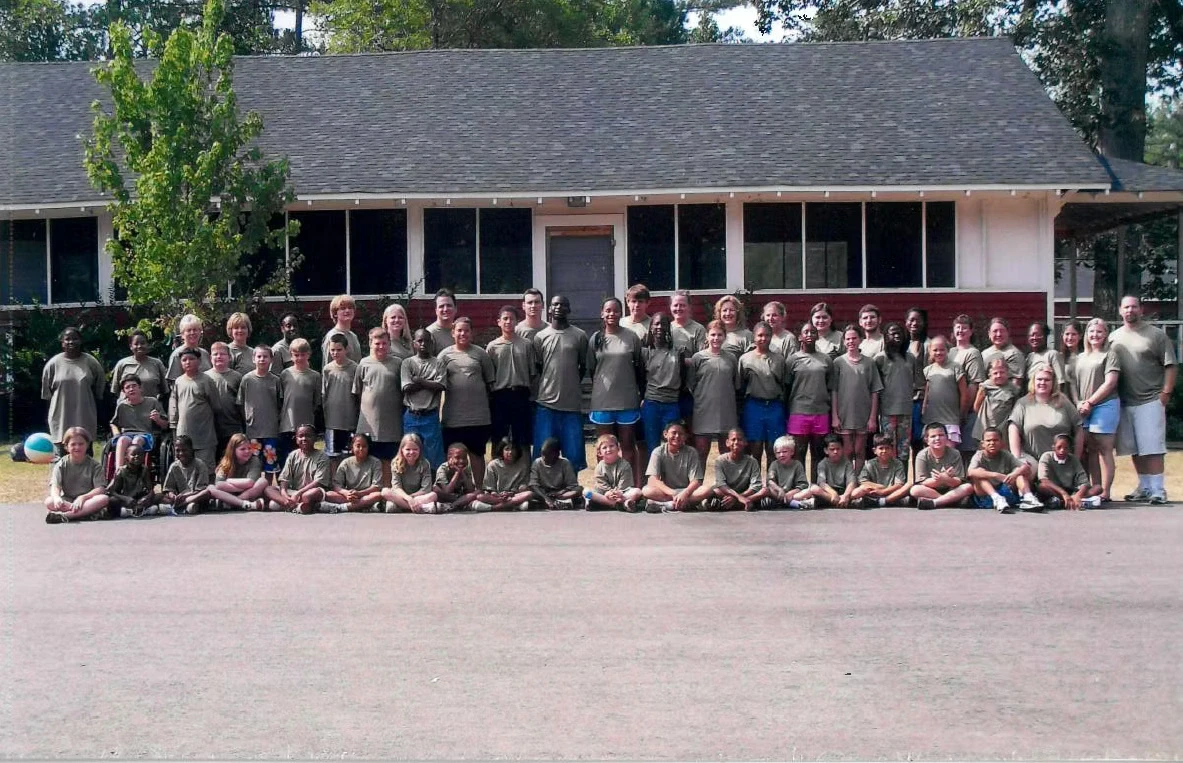 Large group photo of campers and staff in matching olive green shirts sitting and standing in front of a building.