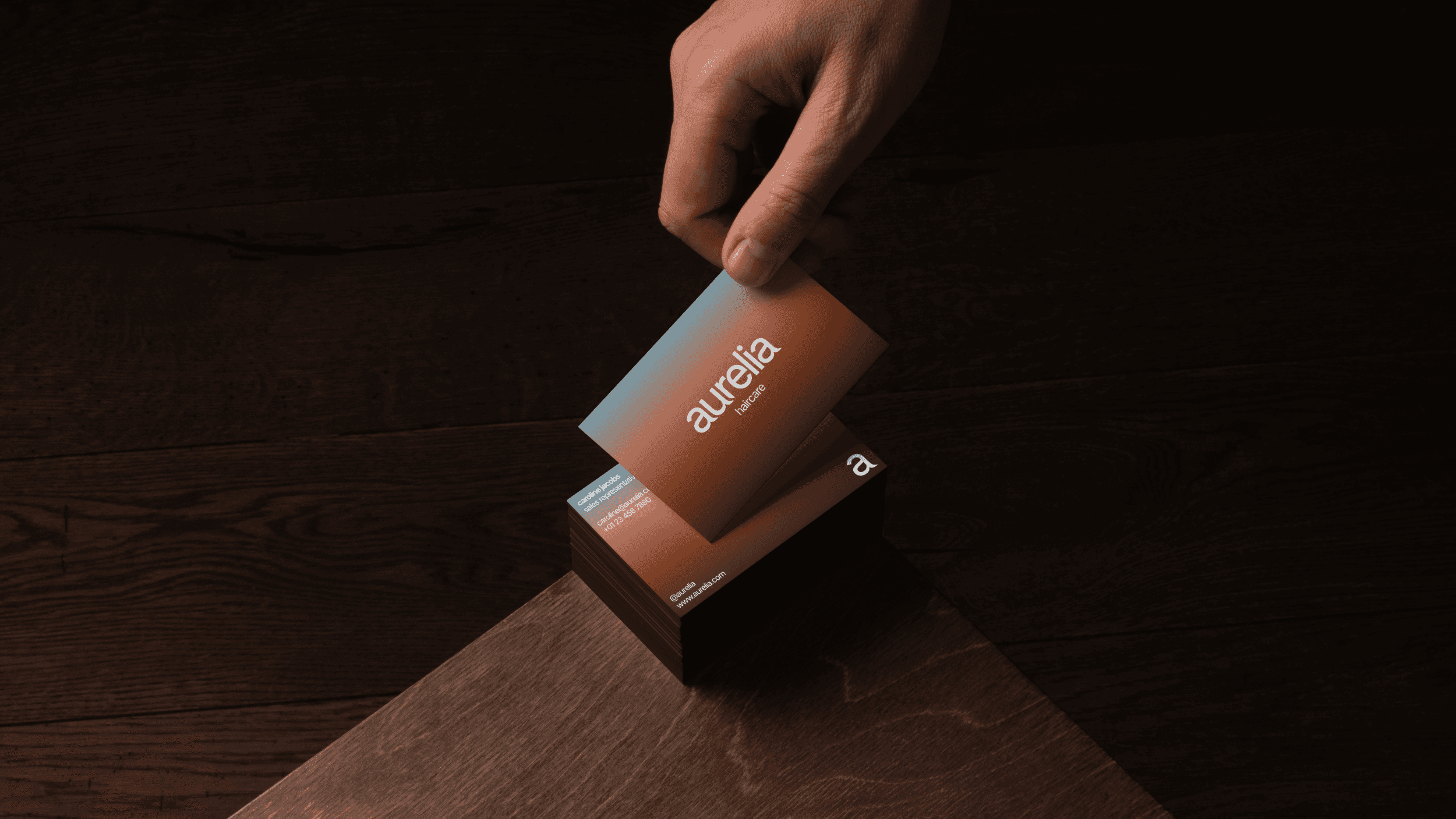 Business cards stacked on the corner of a table with a person's hand holding one of the cards. The floor in the background and the table have a dark wood material. The business cards feature a minimalist design with a blue and brown gradient.