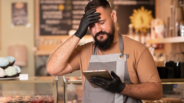 Homem de barba, usando avental e luvas, olhando para um tablet com a mão na testa; ao fundo, balcão com doces.