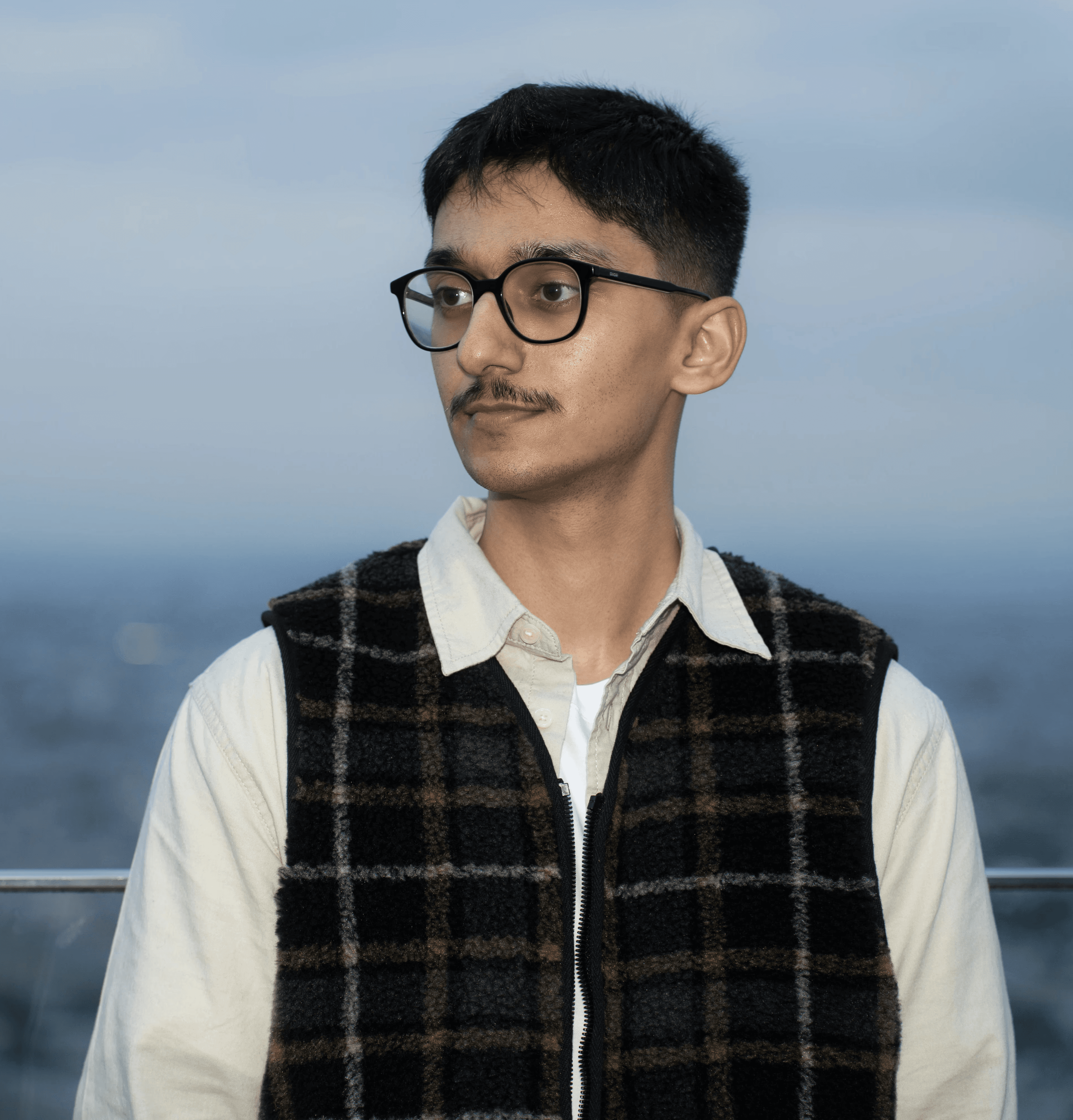 A person wearing glasses and a green jacket smiles while standing on a bridge, with their hands in their pockets.