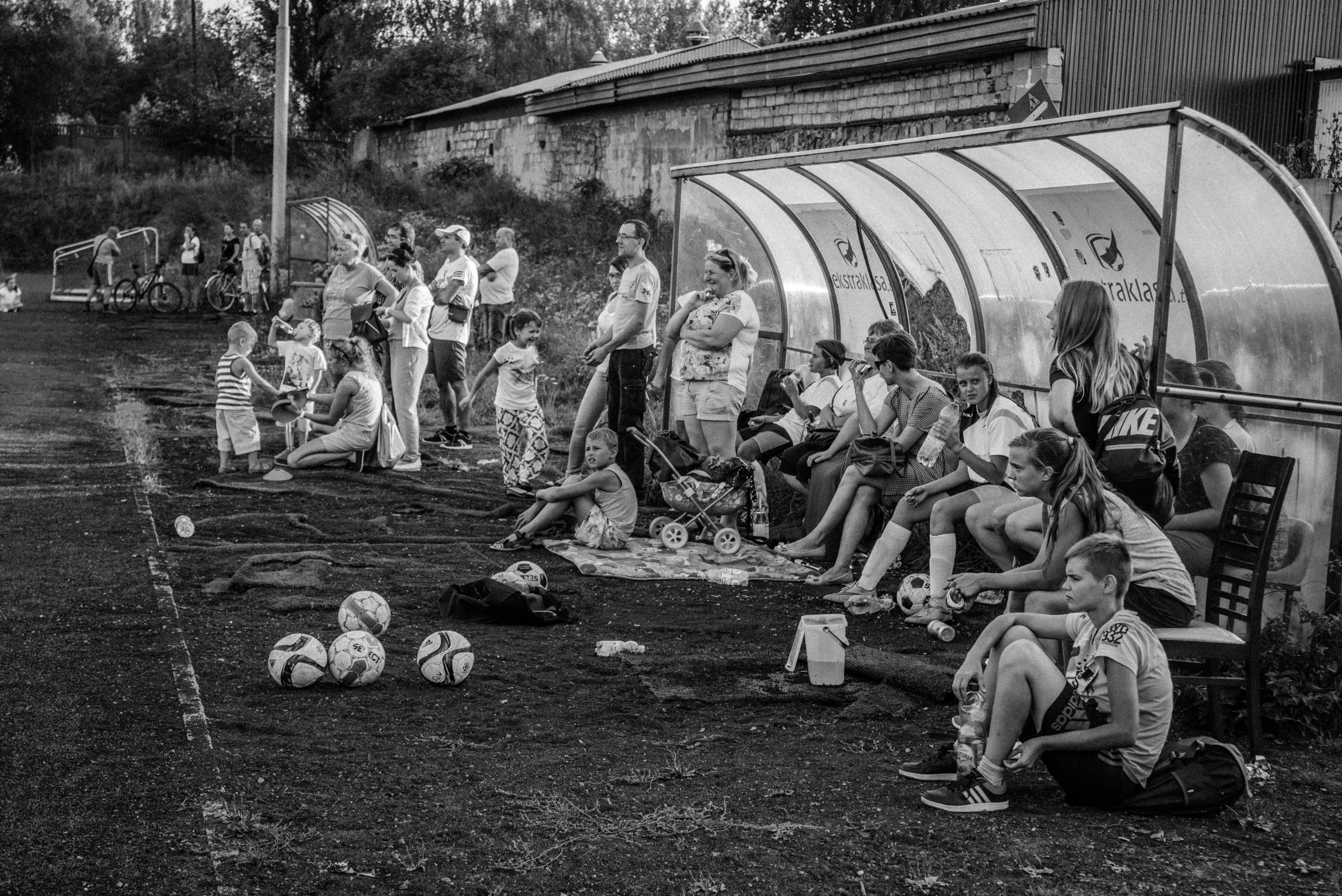 Mecz piłki nożnej. Kibice i piłki leżą na boisku. Ludzie obserwują grę, siedząc na ławkach i stojąc obok.