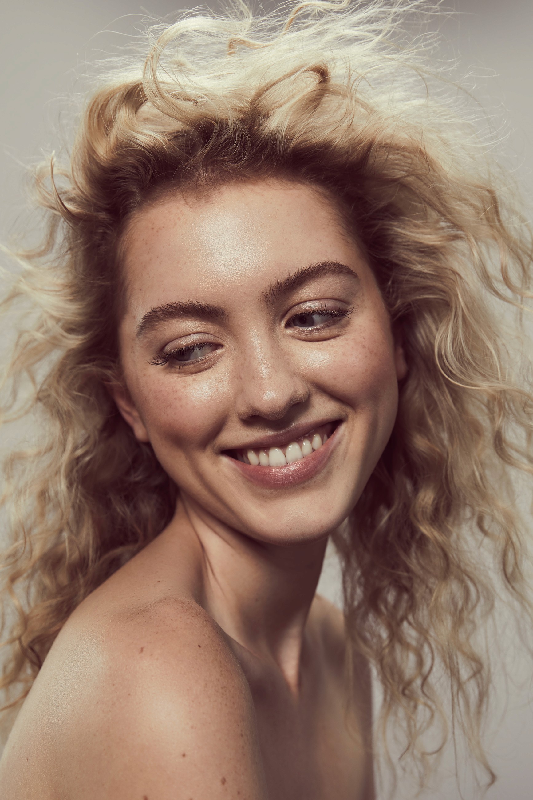 Joyful beauty portrait of woman with windswept blonde curls laughing authentically, highlighting luminous skin, natural freckles, and warm bronze eye makeup in soft studio lighting.