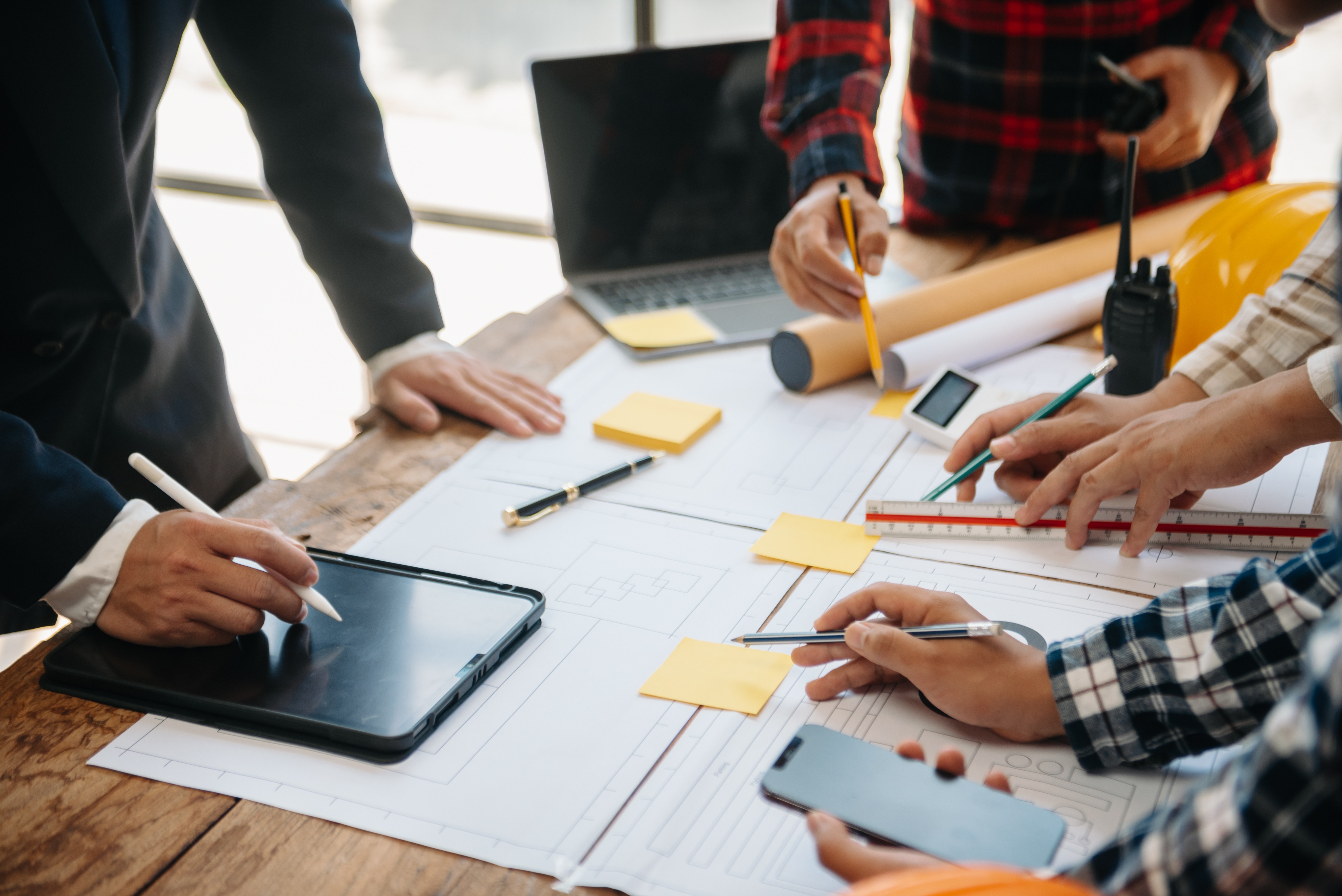 Équipe de professionnels de TCC Especialização collaborant sur un projet architectural ou d'ingénierie, utilisant des tablettes, des ordinateurs portables et des dessins techniques. Réunion de travail axée sur la planification et l'exécution de projets détaillés.