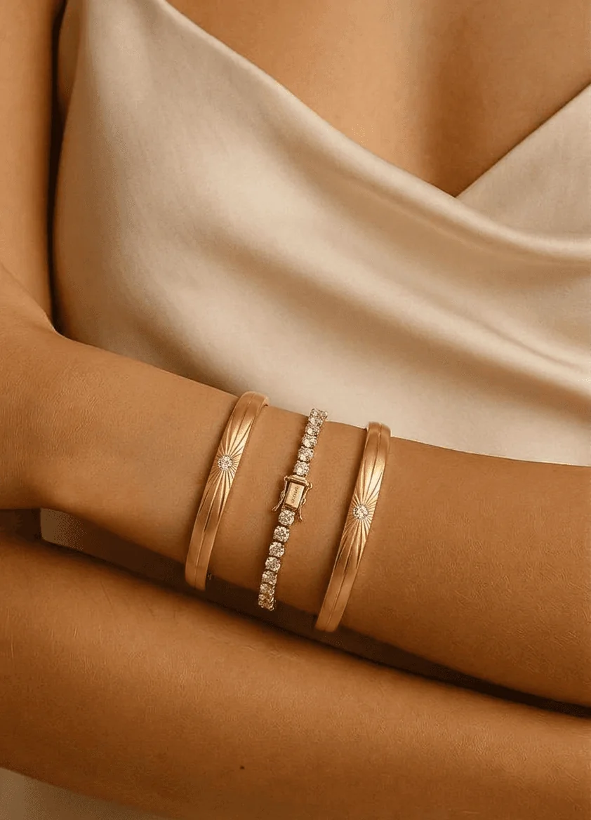 Close-up of a woman's arm wearing three elegant gold bracelets against a soft, draped fabric background.