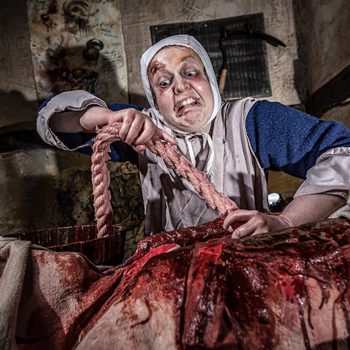 A person in historical attire with a distressed expression holds a bloody rope, over a blood-stained cloth in a dim, eerie room.