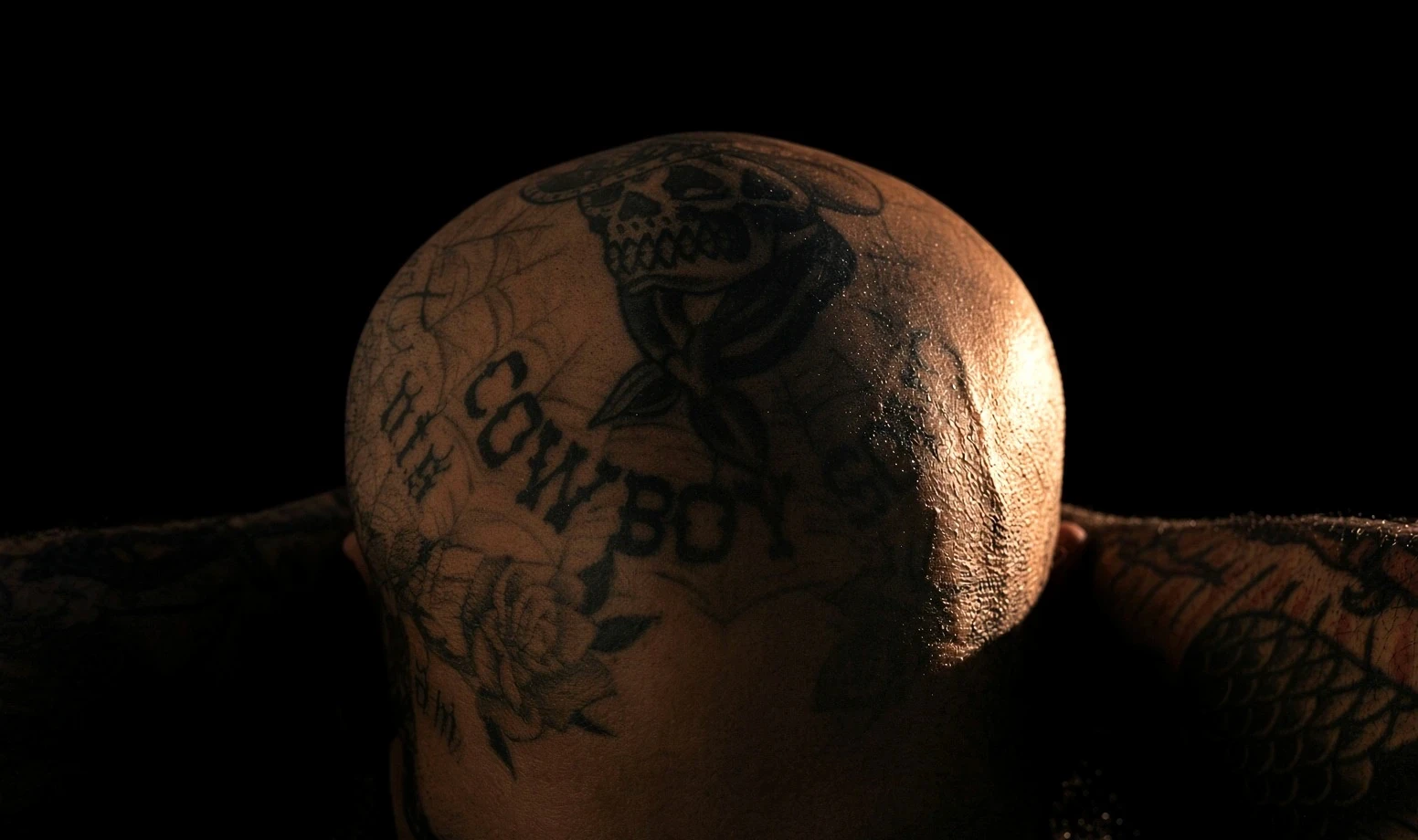 Close-up of Fast Ronald's tattooed head from behind, lit from the side against a black background