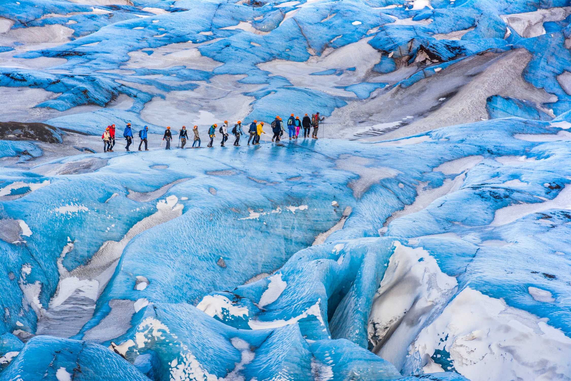 A group of hikers in colorful outdoor gear walks across a vast, textured blue and white glacier under bright daylight.